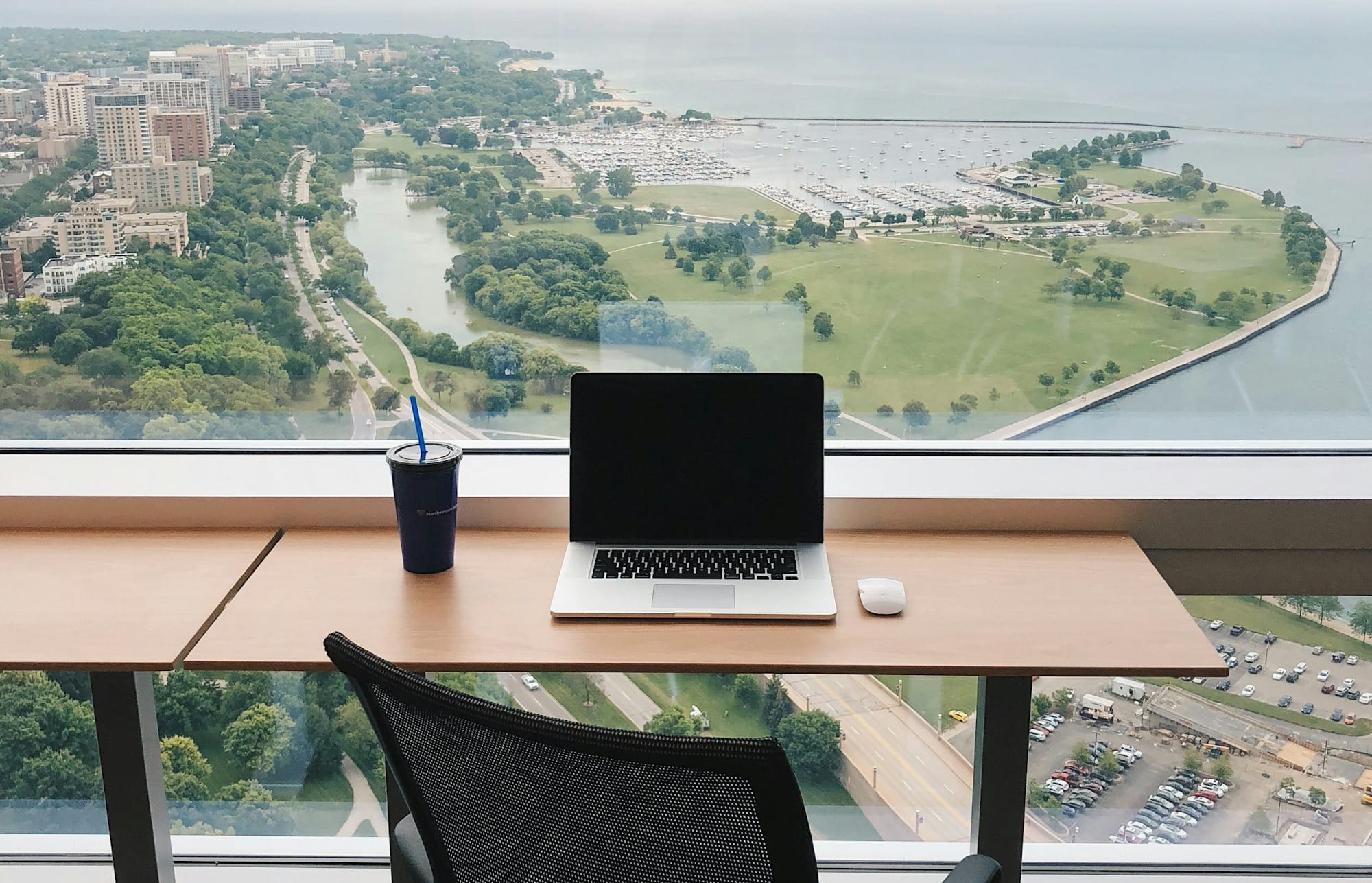 A laptop and mouse with a reusable cup and straw sitting nearby on a desk overlooking a city park as seen from a high-rise w